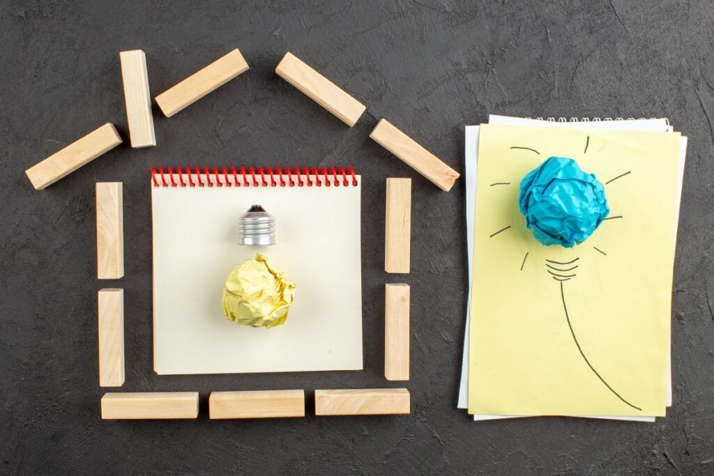 top-view-house-shaped-wood-blocks-goals-written-notepad-idealight-bulb-paper-black